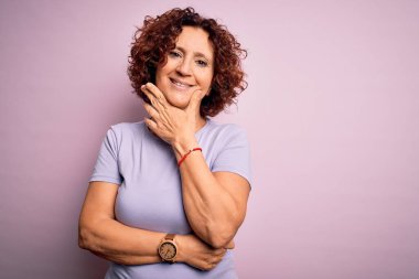 Orta yaşlı güzel kıvırcık saçlı kadın pembe arka plan üzerine günlük tişört giyip kameraya bakarken kolları çapraz ve çenesi havada gülümsüyor. Olumlu düşünüyorum..