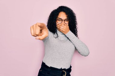 Genç Afro-Amerikan kadın balıkçı yaka kazak ve pembe arka planda gözlük takıyor. Sana gülüyorlar, kamerayı işaret ediyorlar.