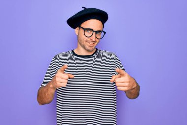 Mor arka plan üzerinde çizgili tişört ve Fransız beresi giyen, mutlu ve komik yüzlü parmakları kameraya gösteren yakışıklı bir adam. İyi enerji ve titreşimler.
