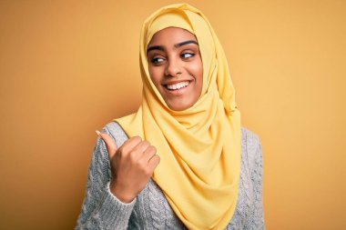 Genç ve güzel Afro-Amerikalı kız, izole edilmiş sarı arka plan üzerinde Müslüman başörtüsü takıyor, gülümsüyor ve başparmağıyla yan tarafa bakıyor..