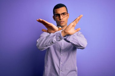 Yakışıklı Afro-Amerikalı adam mor arka planda çizgili gömlek ve gözlük takıyor. Kollarını kavuşturup negatif işaret veriyor, kızgın surat yapıyor.