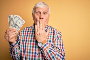 Kıdemli yakışıklı beyaz adam, elinde bir deste banknotla sarı arka planda, ağzı kapalı, utancından ağzı açık, korkunun ifadesi, sessizlikten korkan, gizli bir konsept.