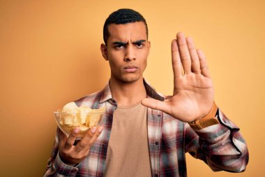 Yakışıklı Afrikalı Amerikalı genç adam elinde patates kızartması ile sarı arka planda dur işareti yapıyor ciddi ve kendinden emin bir ifadeyle, savunma jesti yapıyor.