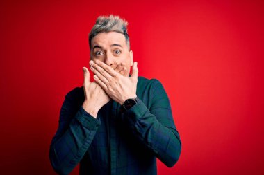 Kırmızı izole arka planda zarif yeşil tişört giyen genç ve modern bir adam yanlışlıkla ağzı ellerle kapatarak şok etti. Gizli konsept.