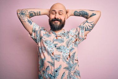 Sakallı ve dövmeli yakışıklı, pembe arka planda rahat ve esneyen, kolları ve elleri başının arkasında mutlu bir şekilde gülümseyen sıradan bir çiçek gömleği giyen kel adam.