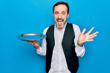 Orta yaşlı, yakışıklı garson, mavi arka planda duran tepsiyi tutarak başarıyı mutlu bir gülümsemeyle ve kazanan yüz ifadesiyle el kaldırarak kutluyor.