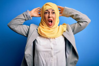 Orta yaşlı esmer iş kadını, geleneksel Müslüman tesettüre bürünmüş mavi arka planda çılgın ve elleri başında korkmuş, şokdan korkan ve ağzı açık biri.