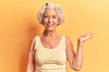 Yaşlı, gri saçlı, günlük kıyafetler giyen neşeli bir şekilde gülümseyen ve avuç içi kameraya bakan bir kadın.. 