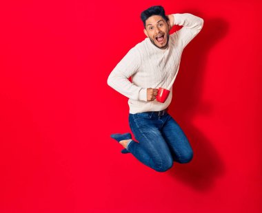 Genç, yakışıklı, gülümseyen, mutlu, bir fincan kahve içen Latin adam. İzole edilmiş kırmızı arka planda yüzünde gülümsemeyle zıplıyor