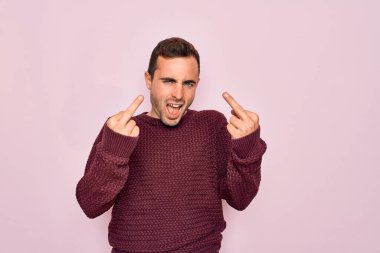 Young handsome man with blue eyes wearing casual sweater standing over pink background Showing middle finger doing fuck you bad expression, provocation and rude attitude. Screaming excited