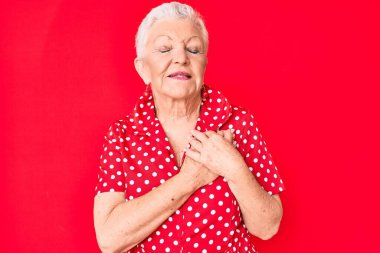 Son sınıf güzeli, mavi gözlü, gri saçlı, kırmızı arka planda günlük giysiler giyen, elleri göğsünde, gözleri kapalı, yüzünde minnet dolu bir jestle gülümseyen bir kadın. sağlık kavramı. 