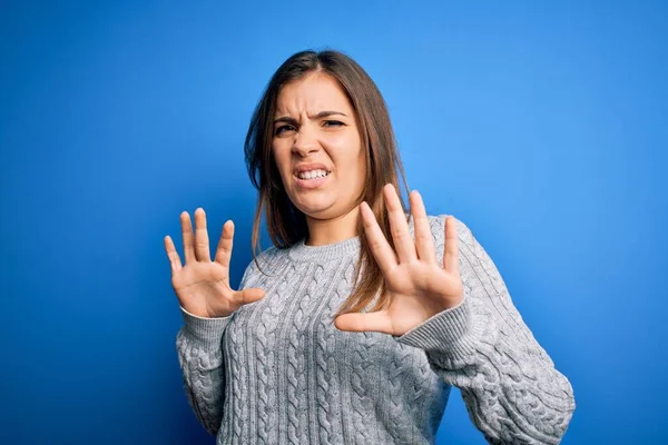 Mavi izole edilmiş arka planda dikilen yünlü süveter giyen genç ve güzel bir kadın korku ifadesinden korkar ve dehşete düşerdi. El hareketlerini durdur, şok içinde bağır. Panik konsepti.