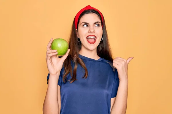 年轻美丽的女孩吃新鲜的有机健康的绿色苹果 背景是黄色的 她用大拇指指着旁边 脸上挂着快乐的笑容 — 图库照片