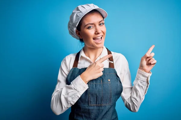 年轻美丽的面包师女人 蓝眼睛 头戴围裙 头戴蓝色背景的帽子 微笑着看着摄像机 双手和手指指向旁边 — 图库照片