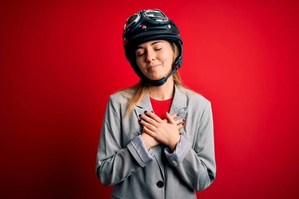 目を閉じて胸に手で笑顔と顔に感謝のジェスチャー赤い背景の上にオートバイのヘルメットを身に着けている若い美しいブロンドのオートバイの女性 健康コンセプト — ストック写真