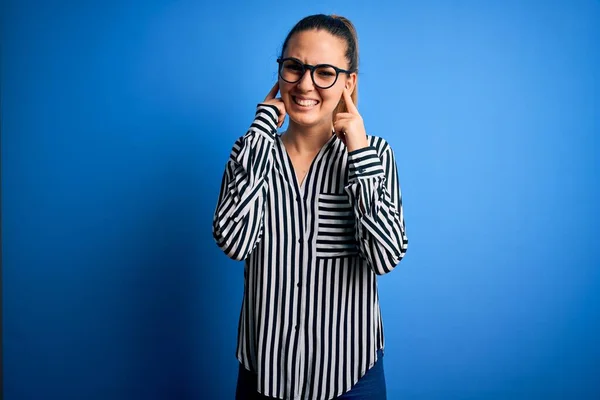 Beautiful blonde woman with blue eyes wearing striped shirt and glasses over blue background covering ears with fingers with annoyed expression for the noise of loud music. Deaf concept.