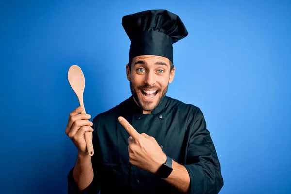 年轻的厨师 留着胡子 身穿制服 蓝色背景上拿着木勺 手指头指指点点 非常高兴 — 图库照片