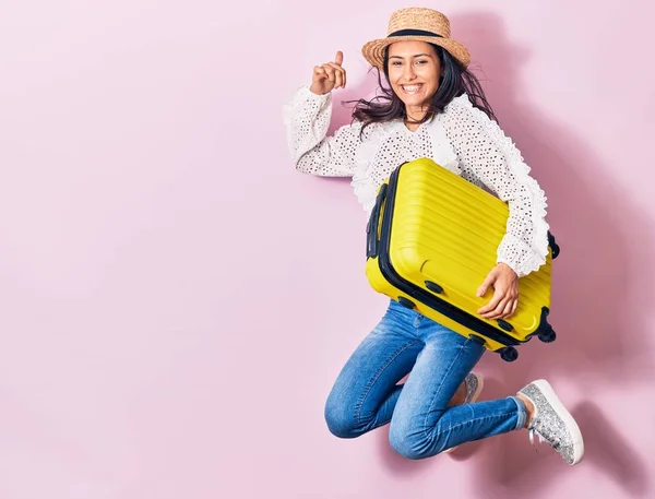 Young beautiful woman on vacation wearing summer clothes and hat smiling happy. Jumping with smile on face holding cabin bag over isolated background