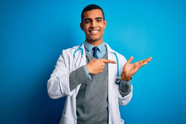 一位英俊的非洲裔美国医生 身穿外套 带着听诊器 头戴蓝色背景眼镜 一边手牵着手 一边指指点点着相机 一边惊讶地微笑着 — 图库照片
