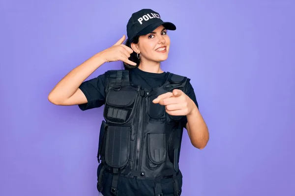 Young police woman wearing security bulletproof vest uniform over purple background smiling doing talking on the telephone gesture and pointing to you. Call me.