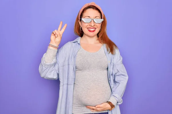 Young redhead pregnant woman expecting baby wearing funny thug life sunglasses smiling with happy face winking at the camera doing victory sign. Number two.