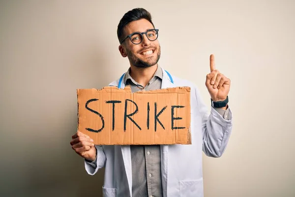 Young handsome doctor man protesting holding cardboard with strike message surprised with an idea or question pointing finger with happy face, number one