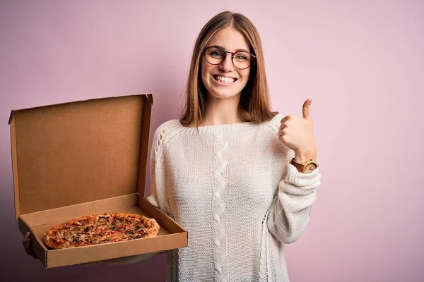 美しい赤毛女性を保持配達ボックスでイタリアの新鮮なピザ以上ピンクの背景幸せなで大きな笑顔を行いますOkサイン 親指アップと指 優れたサイン — ストック写真