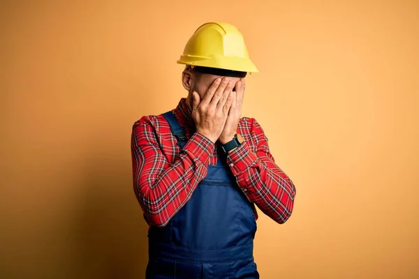 泣きながら手で顔を覆う悲しい表情で黄色の隔離された背景に建設制服と安全ヘルメットを身に着けている若いビルダーの男 うつ病の概念 — ストック写真
