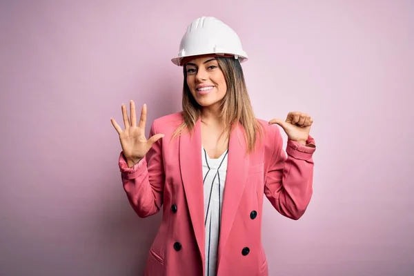 若いです美しいですブルネット建築家女性身に着けています安全ヘルメット上のピンクの背景表示と指差し込み指番号6ながら笑顔自信と幸せ — ストック写真