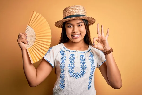 年轻的亚洲旅游姑娘在度假时戴着夏帽 手扇罩在黄色的背景上 手握手签 很好的符号 — 图库照片