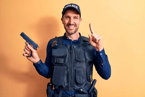 Handsome policeman wearing uniform and bulletprof holding gun over yellow background smiling with an idea or question pointing finger with happy face, number one