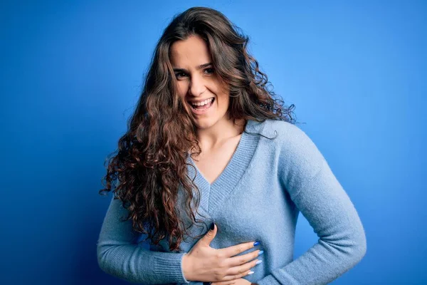 Young beautiful woman with curly hair wearing blue casual sweater over isolated background smiling and laughing hard out loud because funny crazy joke with hands on body.