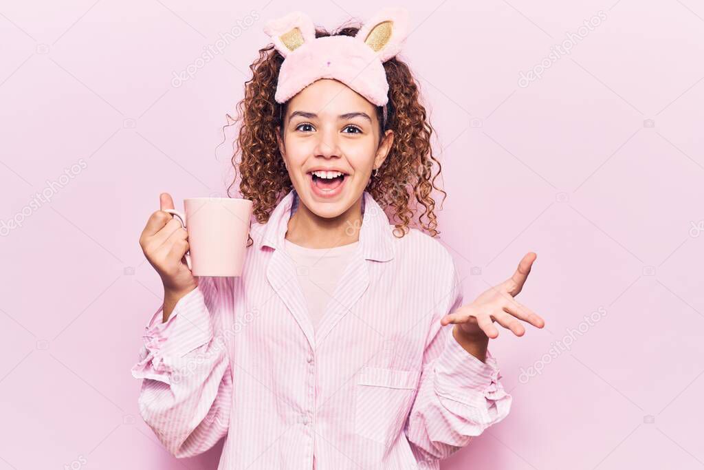 Beautiful kid girl with curly hair wearing sleep mask and pajamas holding coffee celebrating achievement with happy smile and winner expression with raised hand 