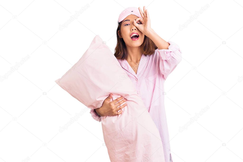 Young beautiful chinese girl wearing sleep mask and pajama holding pillow smiling happy doing ok sign with hand on eye looking through fingers 
