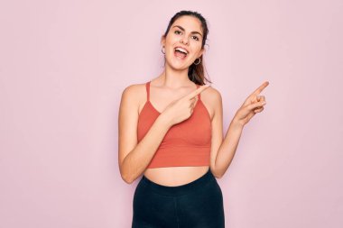 Pembe arka planda spor kıyafetleri giyen, gülümseyen ve kameraya bakan, iki eliyle ve parmaklarıyla işaret eden genç, güzel bir fitness kadını..