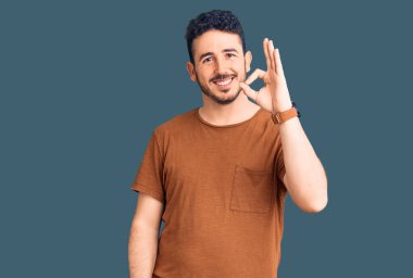 Gündelik kıyafetler giyen genç İspanyol adam gülümsüyor. El ve parmaklarıyla 