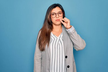 Young hispanic business woman wearing glasses standing over blue isolated background mouth and lips shut as zip with fingers. Secret and silent, taboo talking clipart