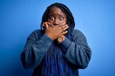 Mavi arka planda günlük süveter giyen Afro-Amerikan kadın ağzı yanlışlıkla elleriyle kapatmış. Gizli konsept.