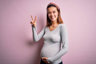 Genç, güzel bir kız hamile, pembe arka planda bebek bekliyor. Gülümsüyor, kameraya göz kırpıyor ve parmaklarıyla zafer işareti yapıyor. İki numara..