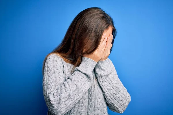 Beautiful young woman wearing casual wool sweater standing over blue isolated background with sad expression covering face with hands while crying. Depression concept.