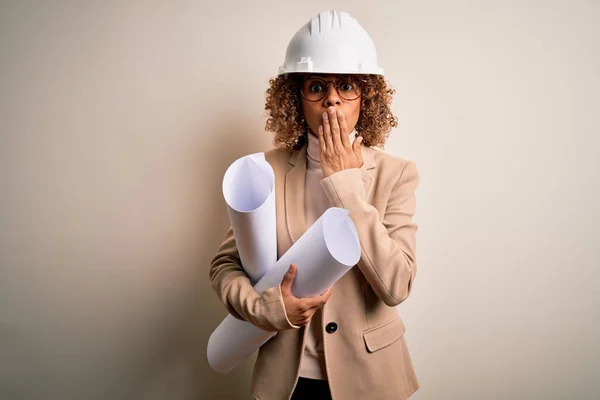 African american curly architect woman wearing safety helmet and glasses holding blueprints cover mouth with hand shocked with shame for mistake, expression of fear, scared in silence, secret concept