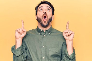 Gündelik giysiler giyen genç İspanyol adam şaşırmış ve şaşırmış bir şekilde yukarı bakıyor ve parmaklarıyla işaret ediyor ve kollarını kaldırıyor.. 