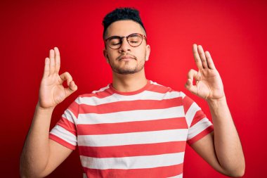 İzole edilmiş kırmızı arka plan üzerine sıradan çizgili tişört ve gözlük takan genç yakışıklı adam rahatlamış ve gözleri kapalı meditasyon yaparken parmaklarıyla gülümsüyor. Yoga konsepti.