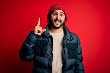 Sakallı, sıradan bir ceket giyen, kırmızı arka planda yün şapkası olan yakışıklı bir genç başarılı bir fikri işaret ediyor. Heyecanlı ve mutlu. Bir numara..