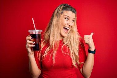 Genç, güzel, sarışın bir kadın kırmızı arka planda saman kullanarak kola içiyor ve gülümseyen bir gülümsemeyle baş parmağıyla onu işaret ediyor.