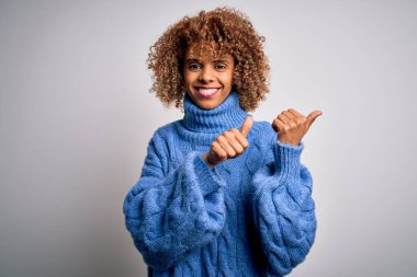 Beyaz arka plan üzerine balıkçı yaka kazak giyen güzel Afro-Amerikalı bir kadın. Arka tarafı işaret ediyor. El ve baş parmakları havada, kendinden emin.