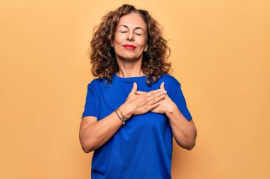 Orta yaşlı, güzel bir kadın, izole edilmiş sarı arka planda dikilmiş tişört giyiyor, elleri göğsünde, gözleri kapalı, yüzünde minnet dolu bir jest ile gülümsüyor. Sağlık kavramı.
