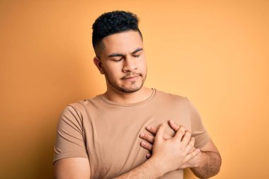 Sarı arka plan üzerinde sıradan tişörtler giyen genç yakışıklı adam elleri göğsünde, gözleri kapalı ve yüzünde minnet dolu bir jest ile gülümsüyor. Sağlık kavramı.