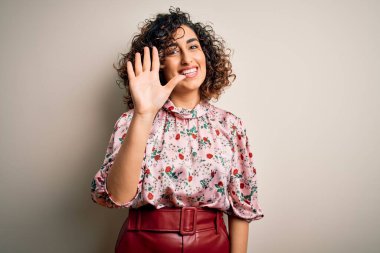 Genç ve güzel kıvırcık Arap kadın çiçekli tişört giyerek izole edilmiş beyaz arka planda duruyor ve parmaklarını 5 numarayı gösterirken kendinden emin ve mutlu gülümsüyor..