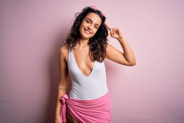 Tatilde kıvırcık saçlı güzel bir kadın pembe arka planda beyaz mayo giyiyor. Tek parmakla kafasını göstererek gülümsüyor. Harika bir fikir ya da düşünce.